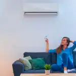 Mujer-joven-feliz-sentada-en-el-sofá-bajo-el-aire-acondicionado-y-ajustando-la-temperatura